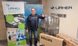 Mika Saari seisoo televisiolaatikoihin nojaten ja näyttää peukaloa, taustalla Urhea-logot